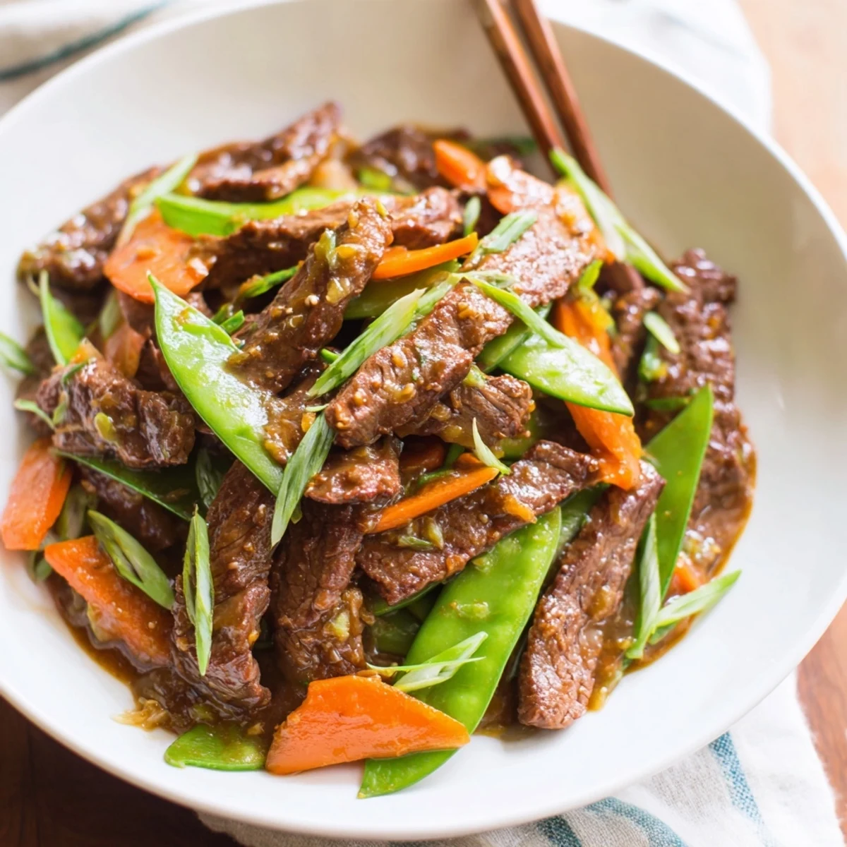 A close-up of Beef Stir Fry with Snow Peas and Carrots beside white rice and chopsticks.