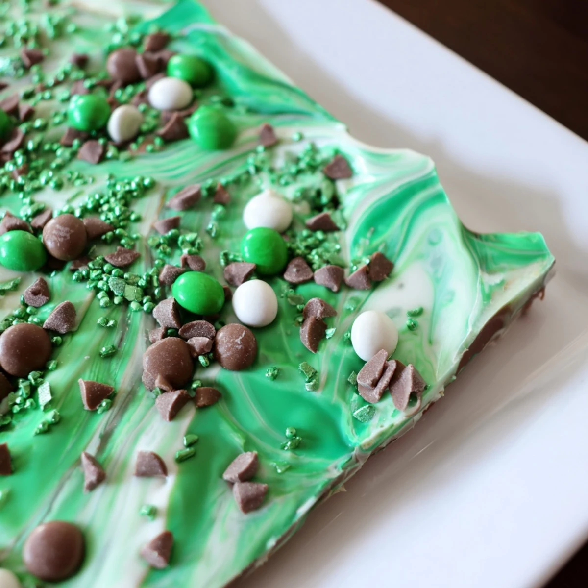 Shiny green St. Patrick's Day Mint Chocolate Bark pieces are arranged on a baking sheet with crushed mint candies and festive sprinkles, perfect for easy holiday snacking.