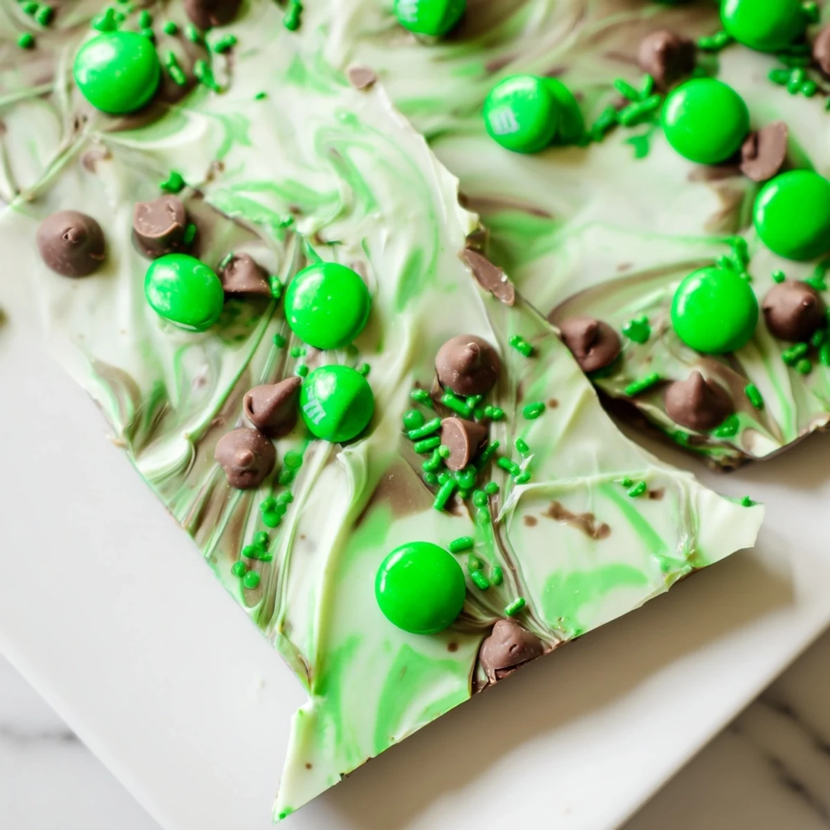 A close-up view shows marbled St. Patrick's Day Mint Chocolate Bark with a minty white chocolate swirl and crunchy green M&Ms, ready to be broken into sharing pieces.