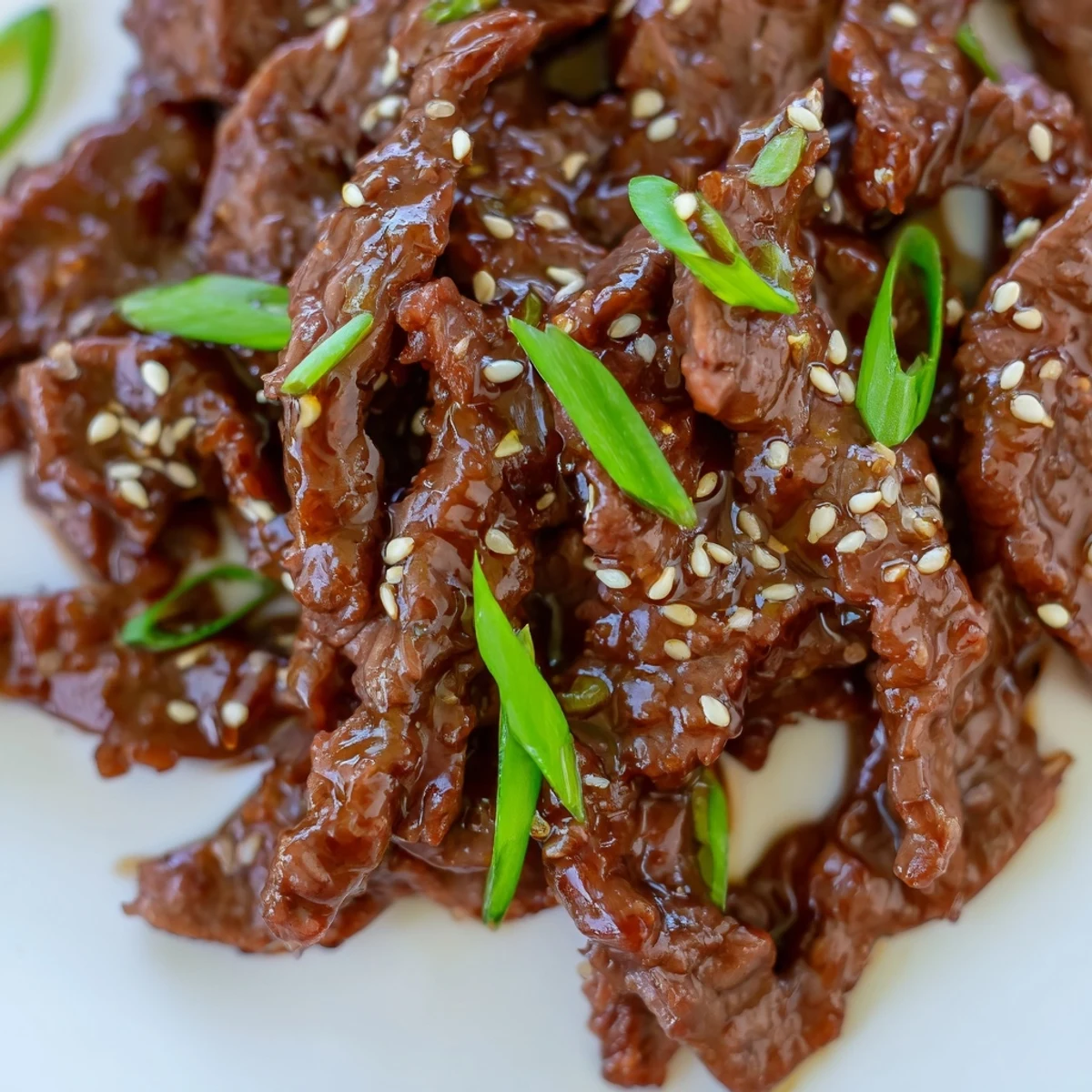 A close-up of Ultimate Slow Cooker Mongolian Beef, tender beef coated in a glossy, savory-sweet sauce, garnished with green onions and sesame seeds served over steamed rice.  