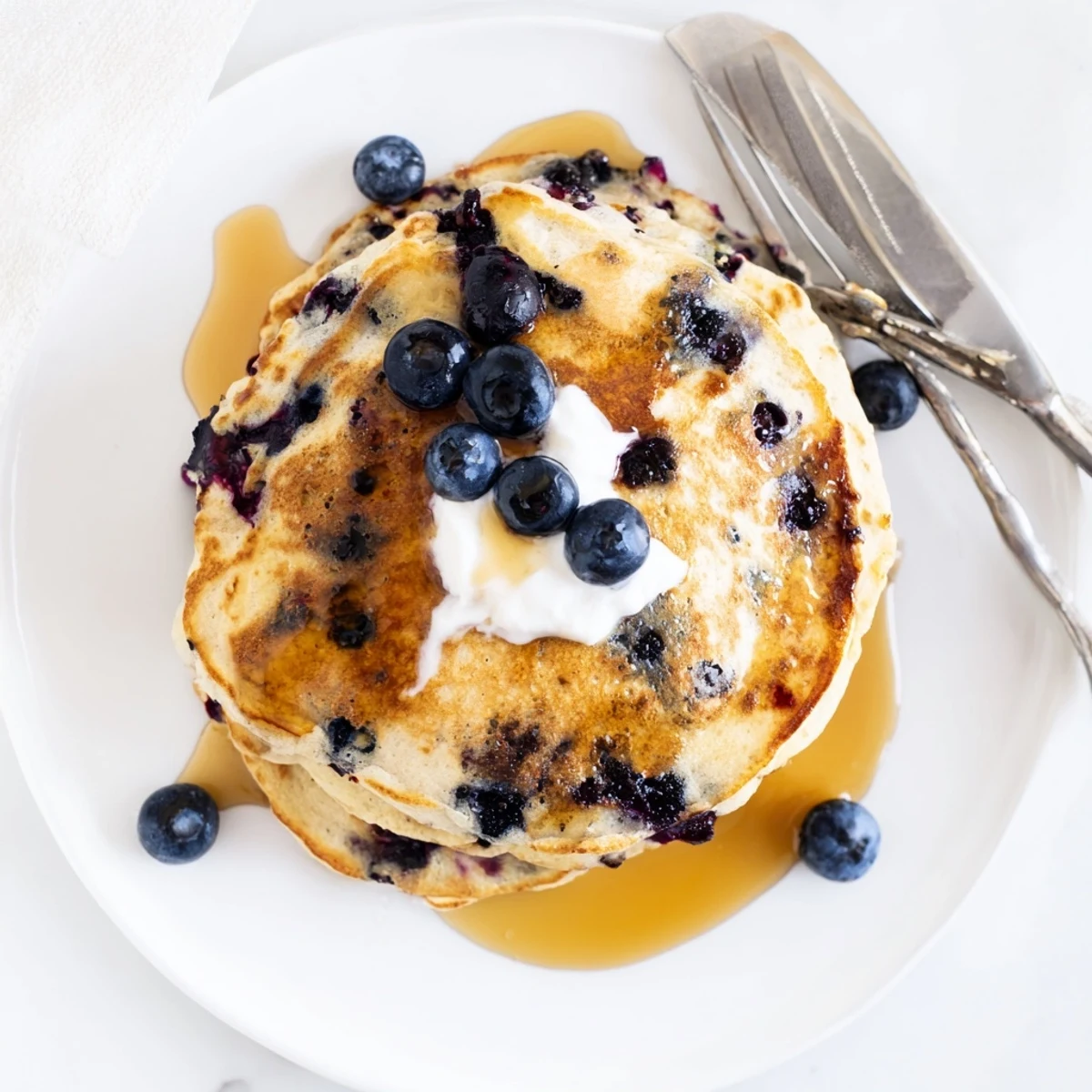 Golden-brown Fluffy Greek Yogurt Blueberry Pancakes stacked high on a plate, drizzled with maple syrup and dusted with powdered sugar. 