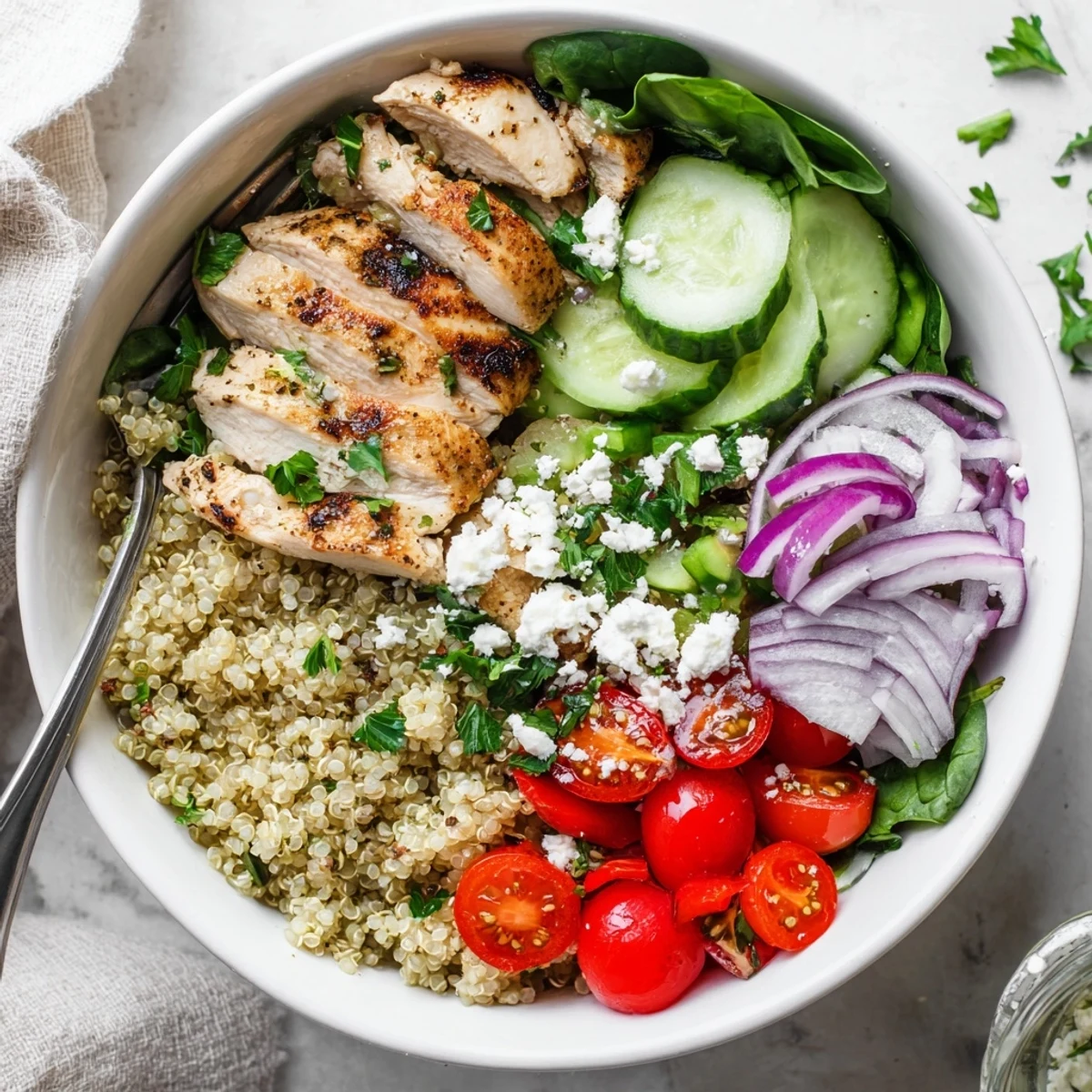Golden-brown grilled chicken slices sit atop fluffy quinoa, vibrant cherry tomatoes, cucumber, red bell pepper, and baby spinach in a white bowl.