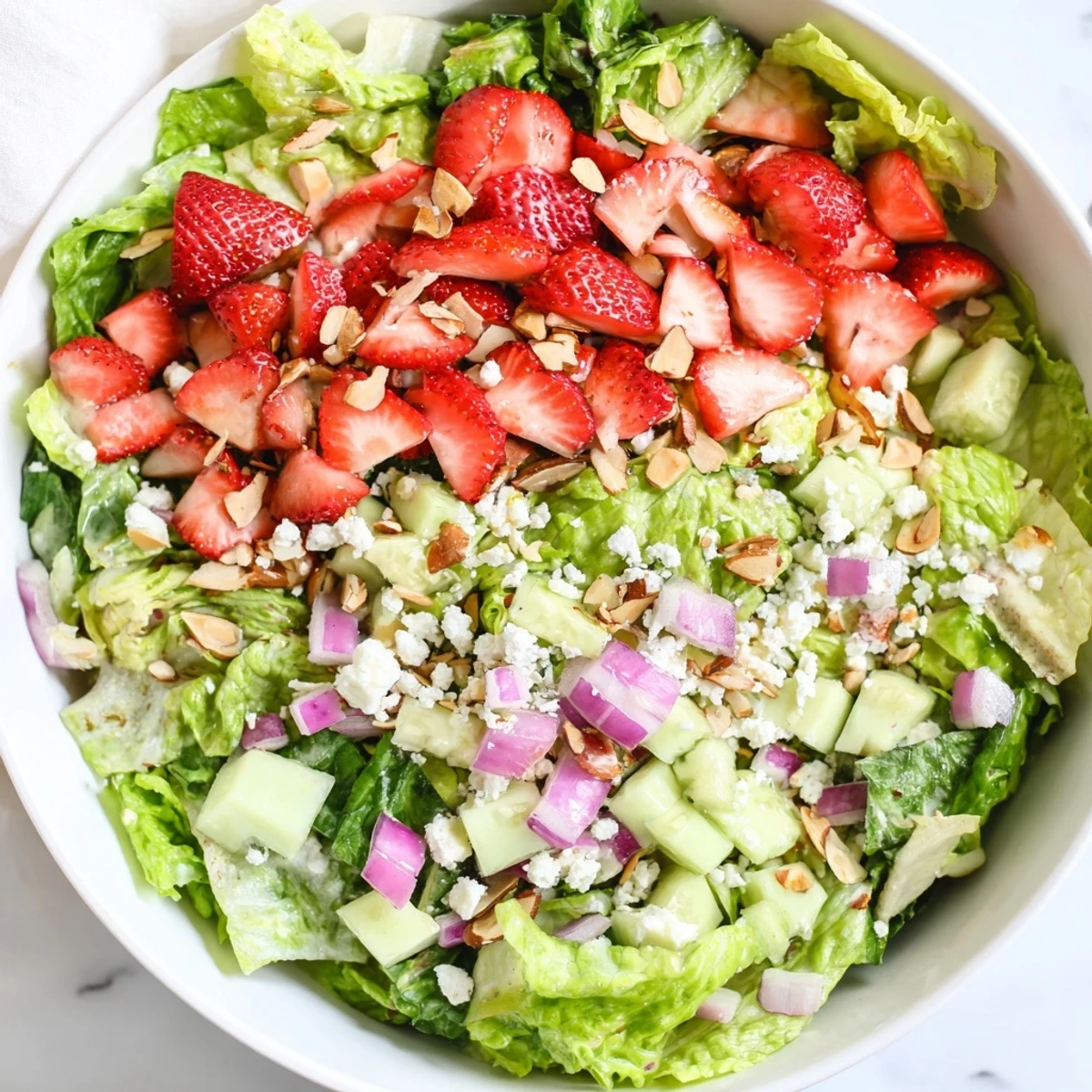 Vibrant bowl of strawberry romaine feta salad featuring sliced strawberries, red onion, cucumber, and crunchy almond toppings drizzled with balsamic dressing.