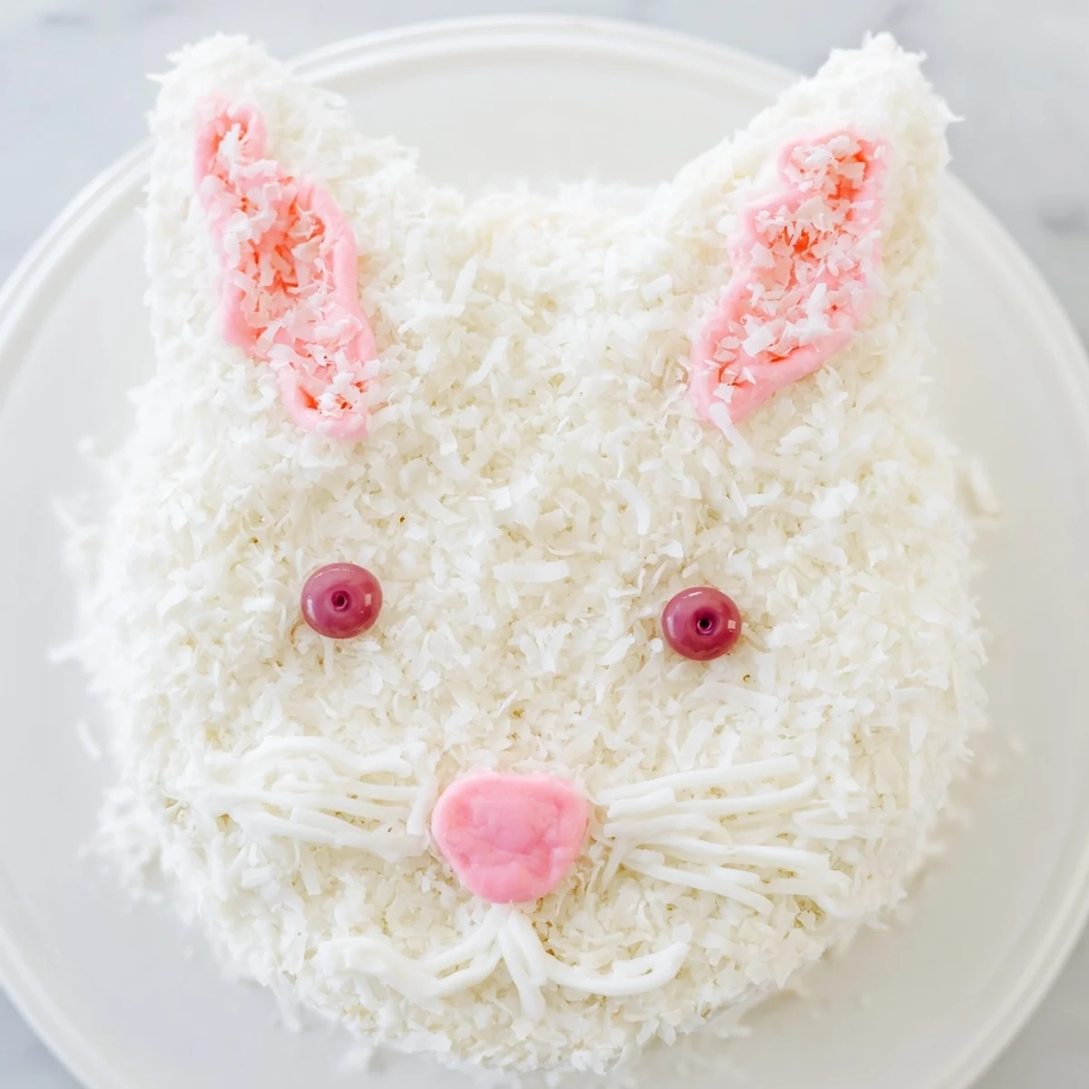 Adorable bunny cake covered in fluffy white coconut frosting with pink candy details and jellybean features